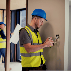 Electrical contractor with blue helmet and vest inspecting an electrical outlet on the wall